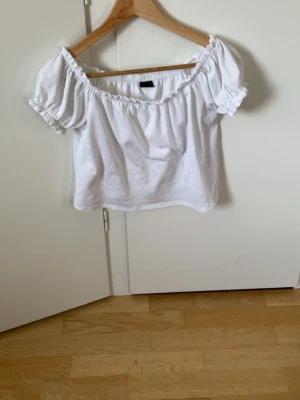 Vit off shoulder topp Gina Tricot - Söt vit off shoulder topp från Gina Tricot med korta puffärmar och resår vid halsringning och ärmslut. Tillverkad i ett lätt, strukturerat tyg som känns luftigt och somrigt. Perfekt för en trendig och avslappnad look.