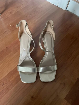 Guldiga sandaletter med blockklack - Snygga sandaletter i metallic guld med bred rem över tårna och smal rem som korsar foten. Skorna har fyrkantig tå och stabil blockklack, samt justerbart spänne vid ankeln. Perfekta för att lyfta vilken outfit som helst.