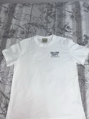Vit t-shirt från Gallery Dept. - Vit t-shirt från Gallery Dept. med svart tryck fram och stort tryck på ryggen där det står 'GALLERY DEPT. HOLLYWOOD, CA.'. Klassisk passform och rund hals. Perfekt för dig som gillar streetwear och enkel stil.