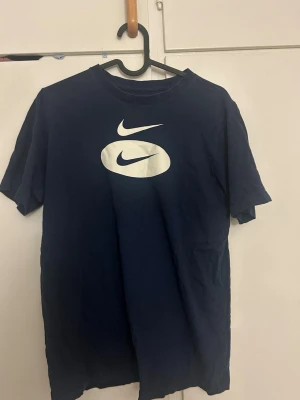 Mörkblå Nike T-shirt med logga - Snygg mörkblå T-shirt från Nike med stor vit logga på bröstet. Klassisk rund hals och korta ärmar. Tillverkad i mjuk bomull som känns skön mot huden. Perfekt för dig som gillar sportig och enkel stil.