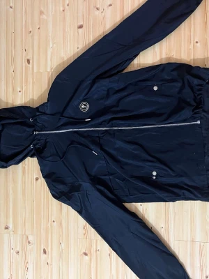 Svart vindjacka med huva - Snygg svart vindjacka med huva och hel dragkedja framtill. Jackan har två fickor med tryckknappar och en liten rund logotyp på bröstet. Lätt material, perfekt för blåsiga dagar. Justerbar nederkant med dragsko.