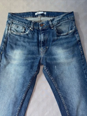 Blå slitna jeans från ZARA - Säljer ett par blå jeans från ZARA med snyggt slitna detaljer och klassisk femficksmodell. Jeansen har en något tvättad look med ljusare partier framtill och bakfickor. Perfekta för en avslappnad och trendig stil.