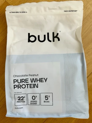 Bulk Pure Whey Protein Choklad/Jordnöt - Nypris 440kr! En påse Bulk Pure Whey Protein med choklad- och jordnötssmak. Innehåller 1 kg pulver, 22 g protein per portion, 0 g tillsatt socker och 5 g BCAA. Förpackningen är vit med svart text och återförslutningsbar topp.