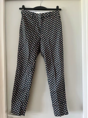 Mönstrad byxdress i svart och beige - Säljer en snygg byxdress med grön bas och beige mönster i rutigt motiv. Byxdressen har raka ben och hög midja, perfekt för dig som gillar statement-plagg. Materialet känns mjukt och följsamt, och den stängs med dragkedja fram.