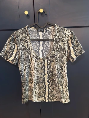 Ormmönstrad topp från Esprit  - Trendig topp från Esprit med ormmönster i beige, svart och grå toner. Modellen har korta ärmar och djup v-ringning. Perfekt för dig som vill sticka ut med en edgy look. Storleken passar till M okså.