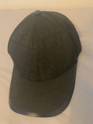 Svart keps med FF Mönster - Snygg svart keps med diskret ton-i-ton mönster över hela ytan. Klassisk böjd skärm och knapp på toppen. Materialet ser ut att vara en blandning av textil och eventuellt lite syntet vid skärmens kant. Perfekt för en stilren och modern look.