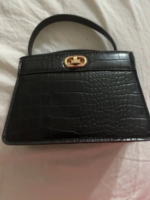 Svart handväska med krokoprägling - Snygg svart handväska i konstläder med krokodilpräglad yta och guldfärgat vridlås. Väskan har en stilren, fyrkantig form och ett kort handtag upptill. Perfekt accessoar för att lyfta din outfit.