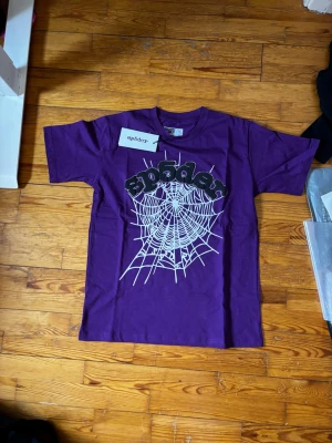 Lila t-shirt med spindelnätsmönster - Säljer en lila t-shirt från sp5der med ett stort vitt spindelnätsmönster och svart text framtill. T-shirten har korta ärmar och en klassisk passform. Perfekt för dig som gillar statement-plagg och coola tryck.