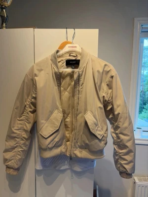 Beige bomberjacka med fickor - Stilren beige bomberjacka med ribbade muddar och krage. Jackan har två snedställda fickor med lock framtill och dragkedja. Quiltad insida för extra komfort. Perfekt för dig som vill ha en clean och enkel look.