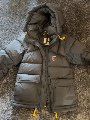 Fjällräven jacka - Snygg mörkgrå dunjacka från Fjällräven med huva och gula detaljer på snörningarna. Jackan har två stora fickor framtill med lock och tryckknappar samt dragkedja och knappslå. Perfekt för kalla vinterdagar. Skulle även kunna tänka mig byta mot en annan färg❤️