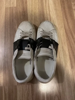 Vita sneakers med svart detalj - Snygga vita sneakers med bred svart läderdetalj över mitten och vita skosnören. Skorna har en klassisk låg profil och en chunky sula. Perfekta för dig som gillar stilrena och moderna sneakers med en twist.