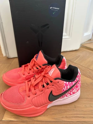 Nike Kobe rosa basketskor - Säljer ett par Nike Kobe basketskor i en stark rosa färg med svart swoosh och vita detaljer. Skorna har mönstrad häl med texten '12:AM' och svart innerfoder. Ovandelen är i mesh och syntetmaterial för bra andningsförmåga och komfort.