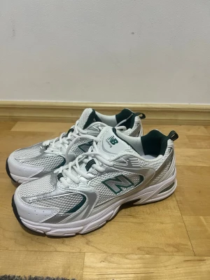 Vita New Balance 530 sneakers - Säljer ett par vita New Balance 530 sneakers med gröna detaljer. Skorna har meshpaneler för extra ventilation och syntetiska partier för stabilitet. Klassisk låg modell med snörning och bekväm sula. Perfekta för en sportig och trendig look.