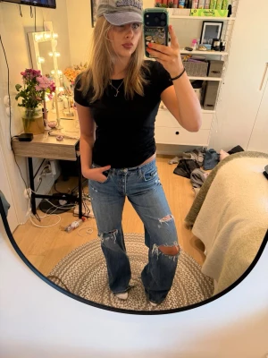 Blå slitna bootcut jeans - Säljer ett par blå bootcut jeans med slitningar och hål på knäna. Jeansen har en klassisk femficksmodell och är tillverkade i denim med en avslappnad passform. Perfekta för en trendig och avslappnad look. Lite slitningar längs ner på jeansen 
