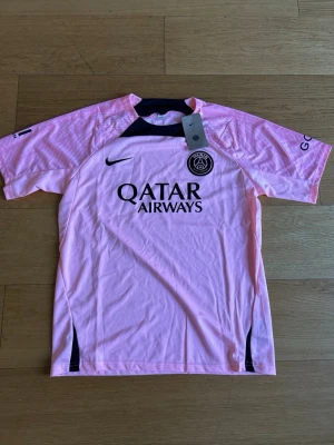 Rosa PSG matchtröja Nike - Snygg rosa PSG matchtröja från Nike med svart logga och detaljer. Framsidan har Qatar Airways-tryck och PSG-emblem på bröstet. Tillverkad i lätt och ventilerande material, perfekt för träning eller match. Korta ärmar och sportig passform.