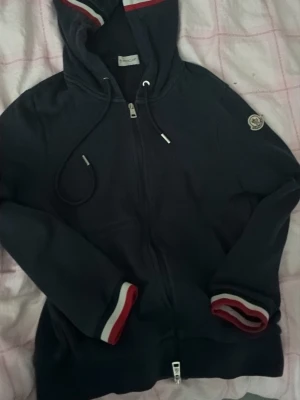 Mörkblå Moncler hoodie med dragkedja - Snygg mörkblå hoodie från Moncler med dragkedja framtill och huva med dragsko. Ärmsluten och huvan har rödvita ränder och Moncler-loggan sitter på ärmen. Perfekt för dig som gillar exklusiva streetwear-plagg. Pris går att diskutera om