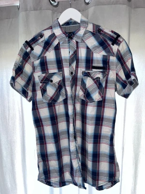 Rutig kortärmad skjorta Jack & Jones - Säljer en snygg kortärmad skjorta från Jack & Jones i rött, blått och vitt rutmönster. Skjortan har två bröstfickor med lock och knapp, axelklaffar och klassisk krage. Perfekt för en avslappnad och trendig stil.