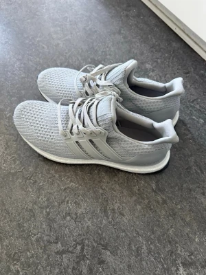 Ljusgrå Adidas sneakers - Säljer ett par ljusgrå Adidas sneakers med stickad ovandel och klassiska tre ränder på sidan. Skorna har vita snören och en bekväm, dämpad sula. Perfekta för dig som gillar stilrena och sportiga skor.