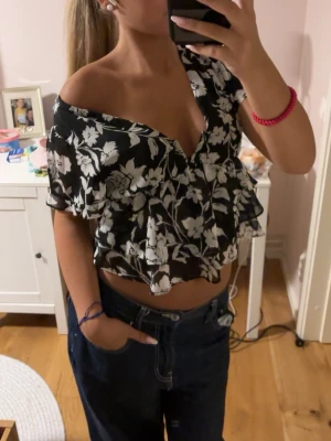 Svart blommig offshoulder topp - Säljer en svart offshoulder topp med vitt blommönster och volangdetaljer. På andra bilden ser man att den är lite sne vid brösten! Annars är den bara provad av mig! Supersnygg top 