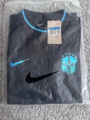 Svart Brasilien Nike fotbollströja - Snygg svart fotbollströja från Nike med Brasilien-emblem på bröstet och blå detaljer vid krage och ärmslut. Tillverkad i lätt och ventilerande material, perfekt för träning eller match. Tröjan har officiella tags kvar och levereras i originalförpackning.