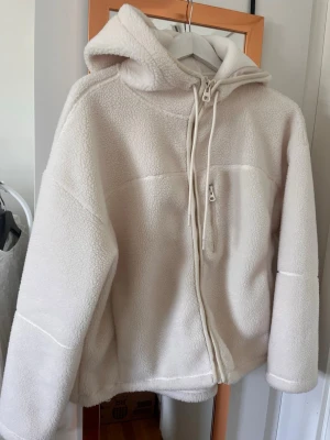 Vit teddyjacka med huva - Mysig teddyjacka i vit med dragkedja framtill och stor huva. Jackan har en avslappnad passform och är tillverkad i fluffigt teddy-material som håller dig varm. Perfekt för kyliga dagar och enkel att matcha med olika outfits. Stor i storleken. 