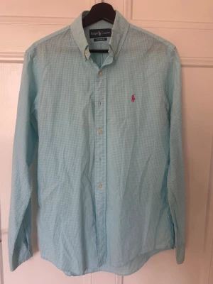Ljusblå rutig skjorta från Ralph Lauren - Snygg ljusblå rutig skjorta från Ralph Lauren med klassisk button-down krage och rosa broderad logga på bröstet. Skjortan har lång ärm och knappar framtill. Perfekt för en clean och stilren look.