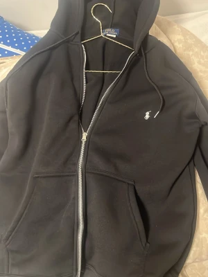 Svart hoodie grisch - Snygg svart hoodie från Polo Ralph Lauren med dragkedja framtill, två fickor och klassisk vit broderad logga på bröstet. Huva med dragsko och mjukt material som känns skönt mot huden. Perfekt för en chill och stilren look.