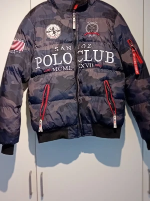 Camouflage dunjacka från Santoz Polo Club - Snygg mörkblå camouflage dunjacka från Santoz Polo Club med stora broderade loggor och text på både fram- och baksida. Jackan har röda dragkedjor, ribbade muddar och flera detaljerade patches. Perfekt för kalla dagarr. Finns defekter, se bilder.