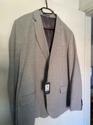 Grå blazer med diskret mönster - Snygg grå blazer med diskret rutmönster och klassisk passform. Kavajen har två framfickor med lock, en bröstficka och knappar vid ärmsluten. Fodrad insida och etikett kvar. Perfekt för dig som vill ha en stilren och tidlös look.