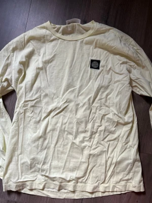 Ljusgul långärmad  Stone Island - Ljusgul långärmad topp från Stone Island med rund hals och ikonisk logopatch på bröstet. Tillverkad i mjuk bomull som känns skön mot huden. Perfekt för dig som gillar stilrena och avslappnade plagg med en exklusiv touch.