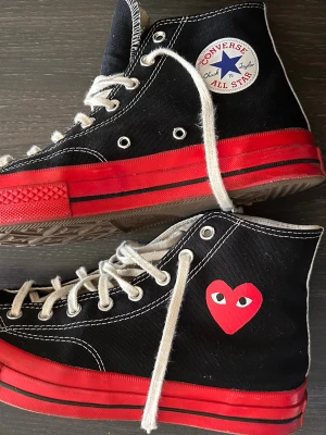 Converse x CDG Play svarta röda highs - Säljer ett par Converse Chuck Taylor All Star 70s i samarbete med Comme des Garçons Play. Svart canvas, röda sulor och det ikoniska CDG-hjärtat på sidan. Vita snören och kontrastsömmar ger en clean look. Perfekta för dig som vill sticka ut.