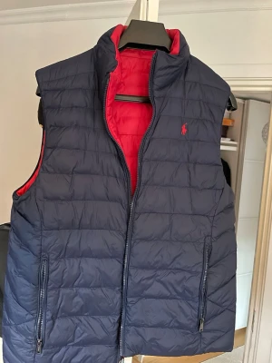 Vändbar dunväst blå/röd Polo Ralph Lauren - Snygg vändbar dunväst från Polo Ralph Lauren i marinblått och rött. Västen har hög krage, två sidofickor med dragkedja och broderad logga på bröstet. Perfekt för lager-på-lager och funkar till många olika stilar. (Barnstorlek XL) (Vanlig storlek XS) Säljer för att jag inte använder den längre. Hör av dig vid några frågor eller intresse!