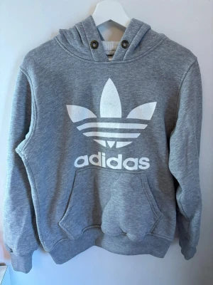 Grå Adidas hoodie - Klassisk grå hoodie från Adidas med stor vit logga på bröstet. Hoodien har magficka, ribbade muddar och huva med snörningshål. Tillverkad i mjuk bomullsblandning, perfekt för en avslappnad look. 