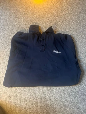 Axel Arigato - Snygg mörkblå pikétröja med krage och knappar framtill. Tröjan har ett diskret broderat textmotiv på bröstet och långa ärmar. Perfekt för en stilren och avslappnad look. Nypris Ca 1800kr