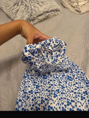 Blommig långklänning med knytband - Söt långklänning med blått blommönster på vit botten. Klänningen har smala axelband med knyt och en rak, smockad överdel. Perfekt för sommarens varma dagar och har en lätt och luftig känsla.