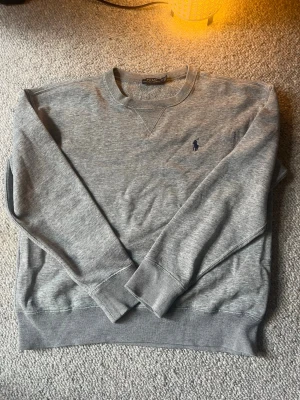 Grå sweatshirt från Polo Ralph Lauren - Nypris ca 1200kr. Klassisk grå sweatshirt från Polo Ralph Lauren med rund halsringning och ribbade muddar. Tröjan har en diskret broderad logga i marinblått på bröstet och är tillverkad i mjuk bomullsblandning. Perfekt för en avslappnad och stilren look.