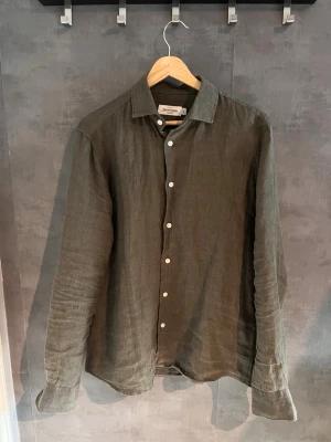 Olivgrön linneskjorta från Dressmann - Säljer en stilren olivgrön skjorta från Dressmann i svalt linne. Skjortan har klassisk krage, vita knappar framtill och långa ärmar. Perfekt för dig som gillar en avslappnad men ändå snygg look.