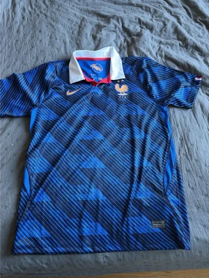Frankrikes vm tröja - Snygg blå fotbollströja från Nike med Dri-FIT-material som håller dig torr. Tröjan har korta ärmar, grafiskt mönster i olika blå nyanser och tryck i gult framtill med texten 'Nos différences nous unissent'. Perfekt för träning eller match.