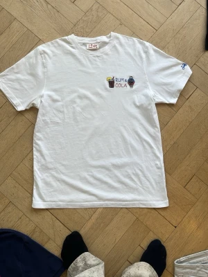 Vit t-shirt med broderi från Saint Barth  - Vit t-shirt från South Bitch med broderat motiv på bröstet som föreställer en drink och en klocka samt texten 'Rum & Cola'. Klassisk passform och rund hals. Perfekt för dig som gillar unika detaljer och chill vibe.