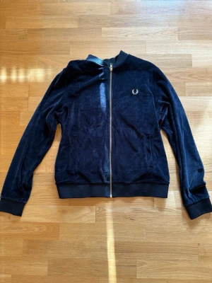 Mörkblå velourtröja från Fred Perry - Mjuk och stilren mörkblå velourtröja från Fred Perry med dragkedja framtill och guldfärgad logga broderad på bröstet. Tröjan har ribbade muddar vid ärmslut och nederkant samt två fickor fram. 