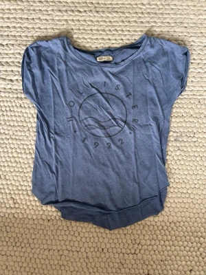 Blå t-shirt från Hollister - Blå t-shirt från Hollister med rund halsringning och tryck framtill med vågmotiv och texten 'Hollister 1922'. Skön passform och tillverkad i mjuk bomull, perfekt för en avslappnad stil.
