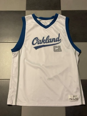Vit basketlinne Oakland CA - Snyggt vitt basketlinne med blåa detaljer och texten 'Oakland CA' på framsidan. På baksidan står det 'California' och siffran 98 i stora grå bokstäver. Perfekt för träning eller streetwear. Tillverkad i ett lätt och ventilerande material.