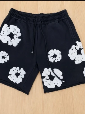 Svarta shorts med vita blommor - Säljer ett par svarta shorts med elastisk midja och dragsko. Shortsen har flera vita blommönster tryckta över hela plagget och är tillverkade i ett mjukt, troligtvis bomullsmaterial. Perfekta för en avslappnad och trendig stil.