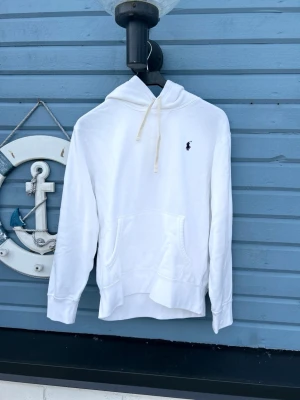 Vit hoodie med logga från Polo Ralph Lauren - Snygg vit hoodie från Polo Ralph Lauren med klassisk marinblå logga på bröstet. Hoodien har huva med dragsko och en stor magficka framtill. Tillverkad i mjukt material och har ribbade muddar vid ärmar och nederkant. Perfekt för en clean och avslappnad stil.