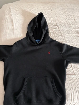 Polo Ralph Lauren Hoodie - 168-176 cm - Snygg svart hoodie från Polo Ralph Lauren med klassisk röd logga broderad på bröstet. Hoodien har en huva och magficka framtill.  Storlek: Youth XL (18–20), motsvarar 168–176 cm. Passar ungefär som XS herr. Perfekt skick.