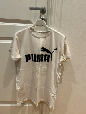 Vit t-shirt med svart Puma-logga - Snygg vit t-shirt från Puma med stor svart logga och puma-tryck på bröstet. Klassisk rund halsringning och korta ärmar. Perfekt till jeans eller shorts för en avslappnad look.