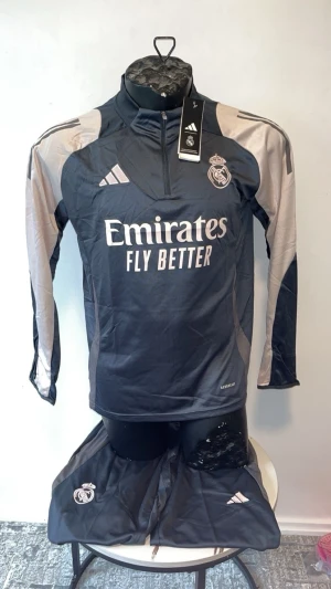 Real Madrid träningsset svart/grå - Snyggt träningsset från Adidas för Real Madrid med långärmad tröja och byxor. Setet är svart med grå detaljer och har klubbmärke samt Adidas-logga på både tröja och byxor. Materialet är lätt och andas, perfekt för träning eller uppvärmning.
