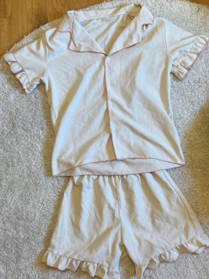 Ljusrosa pyjamas med volangdetaljer - Söt tvådelad pyjamas i ljusrosa med kortärmad skjorta och shorts. Båda delarna har volangdetaljer vid ärm- och benslut samt rosa kontrastsömmar. Skjortan har knäppning framtill och klassisk krage. Perfekt för mysiga kvällar hemma.