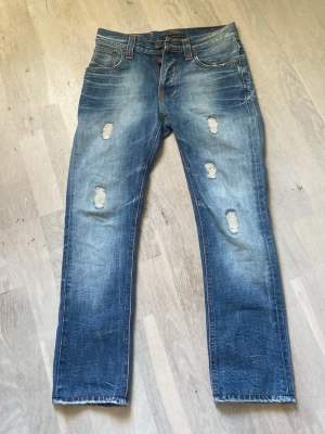 Blå slitna jeans med raka ben - Säljer ett par blå jeans med raka ben och tydliga slitningar framtill. Jeansen har klassisk femficksdesign, orangea sömmar och en snygg tvättad look. Perfekta för dig som gillar en avslappnad och trendig stil.