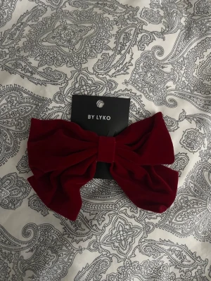 Vinröd sammetsrosett från Lyko - Snygg vinröd hårrosett i sammetsmaterial. Rosetten är stor och fästs enkelt med en metallklämma på baksidan. Perfekt för att ge frisyren en extra touch och passar till många olika looks.